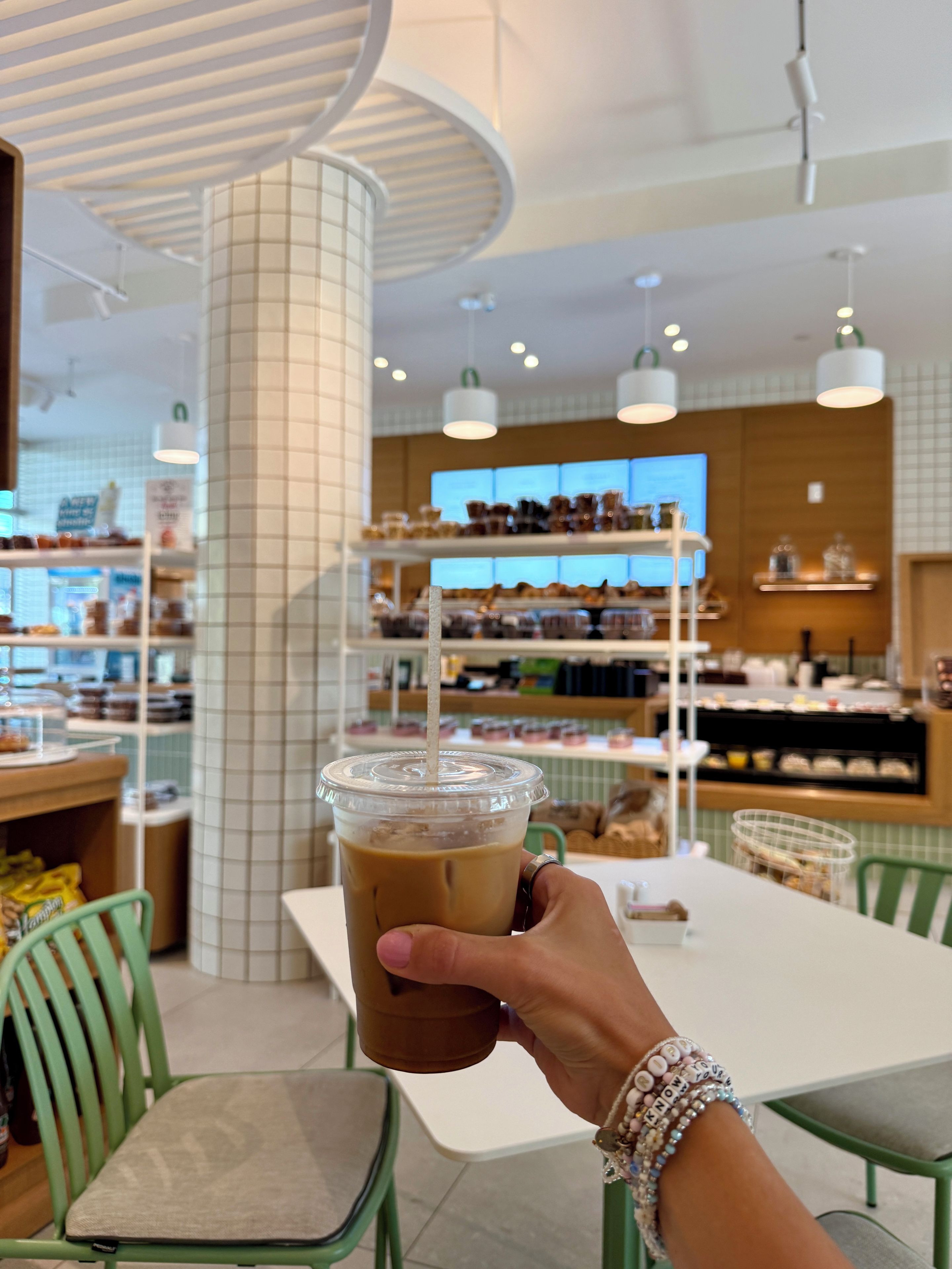 Una persona sosteniendo una taza de café helado con una pajita en una cafetería con pared de azulejos y estantes llenos de pasteles al fondo.