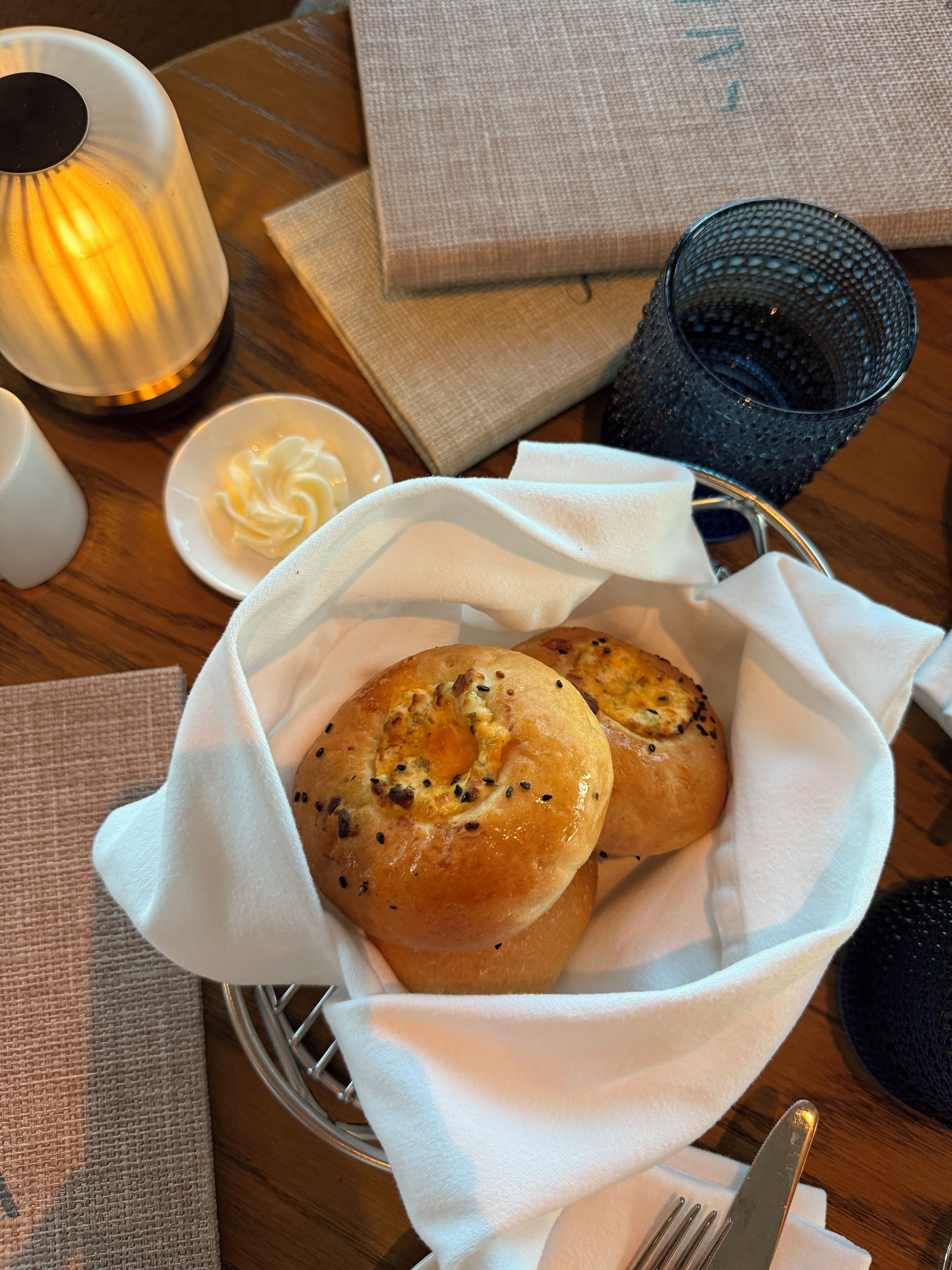 A basket of bread rolls on a table with a candle, a glass, and a small dish of butter.