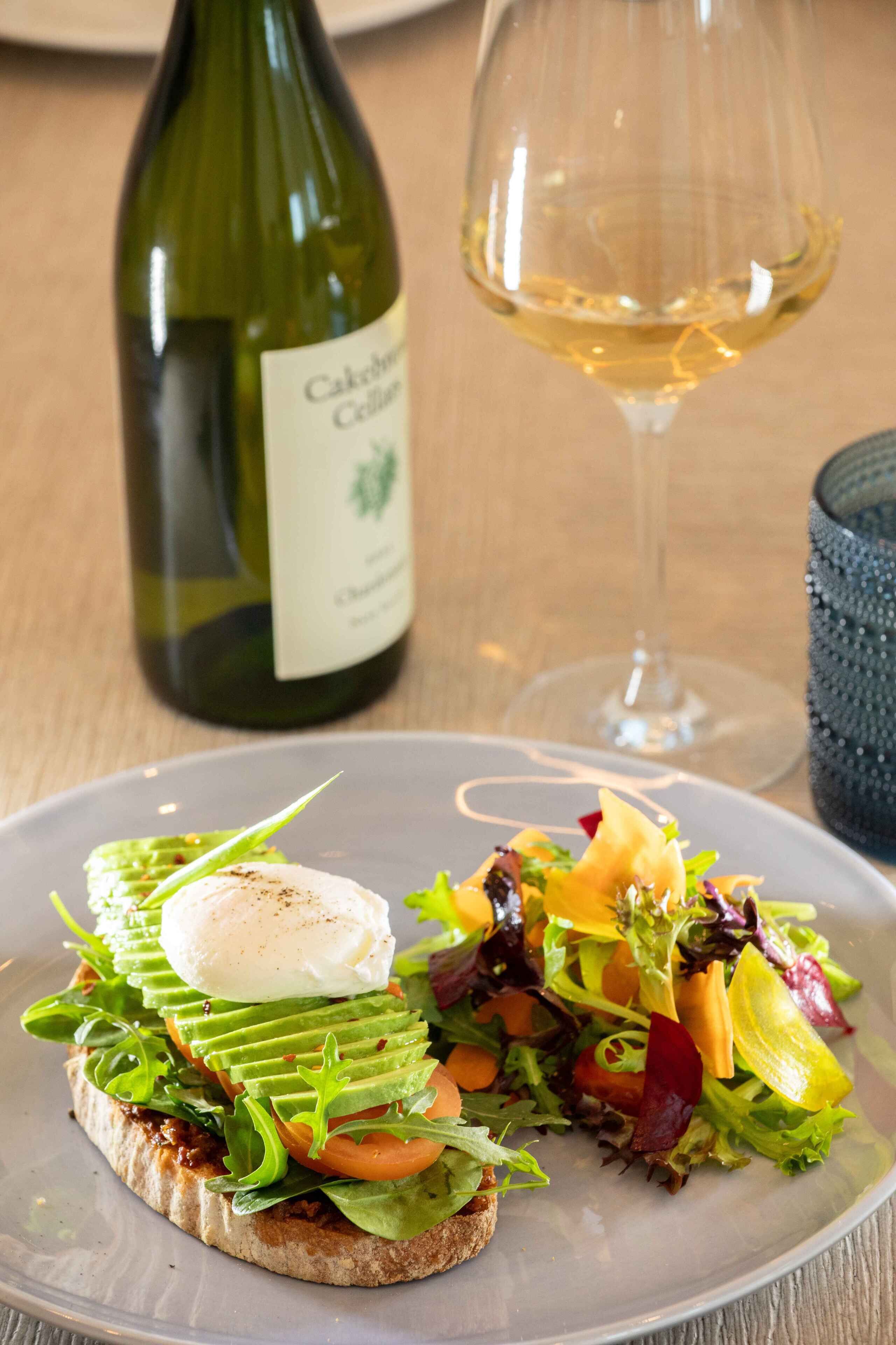 Um prato com salada e ovo pochê em torradas, uma garrafa de vinho e um copo de vinho sobre uma mesa de madeira.