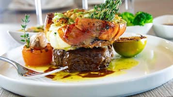 A plate with a steak topped with bacon, garnished with herbs, and accompanied by a lemon and a small tomato.