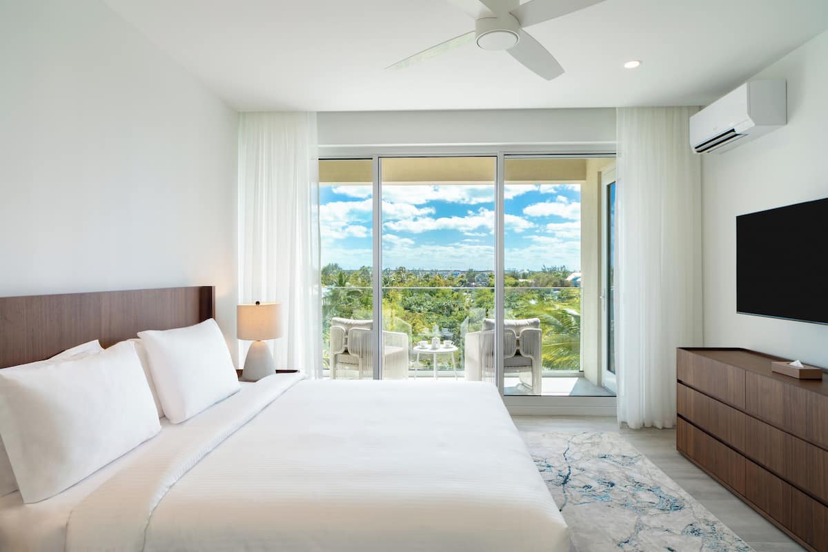 A hotel room with a large bed, a TV, a balcony with chairs, and a view of the outdoors.