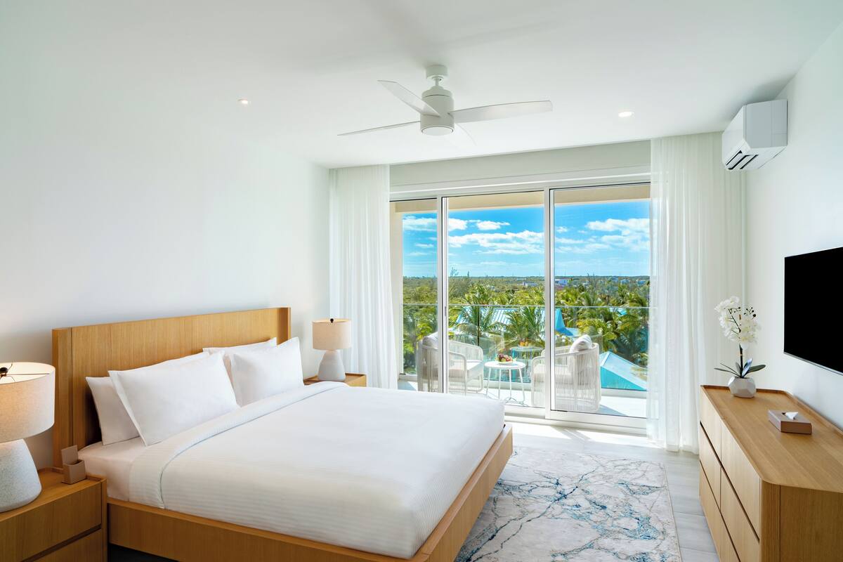 A hotel room with a large bed, a ceiling fan, a TV, a wooden dresser, and a balcony with a view of the ocean.