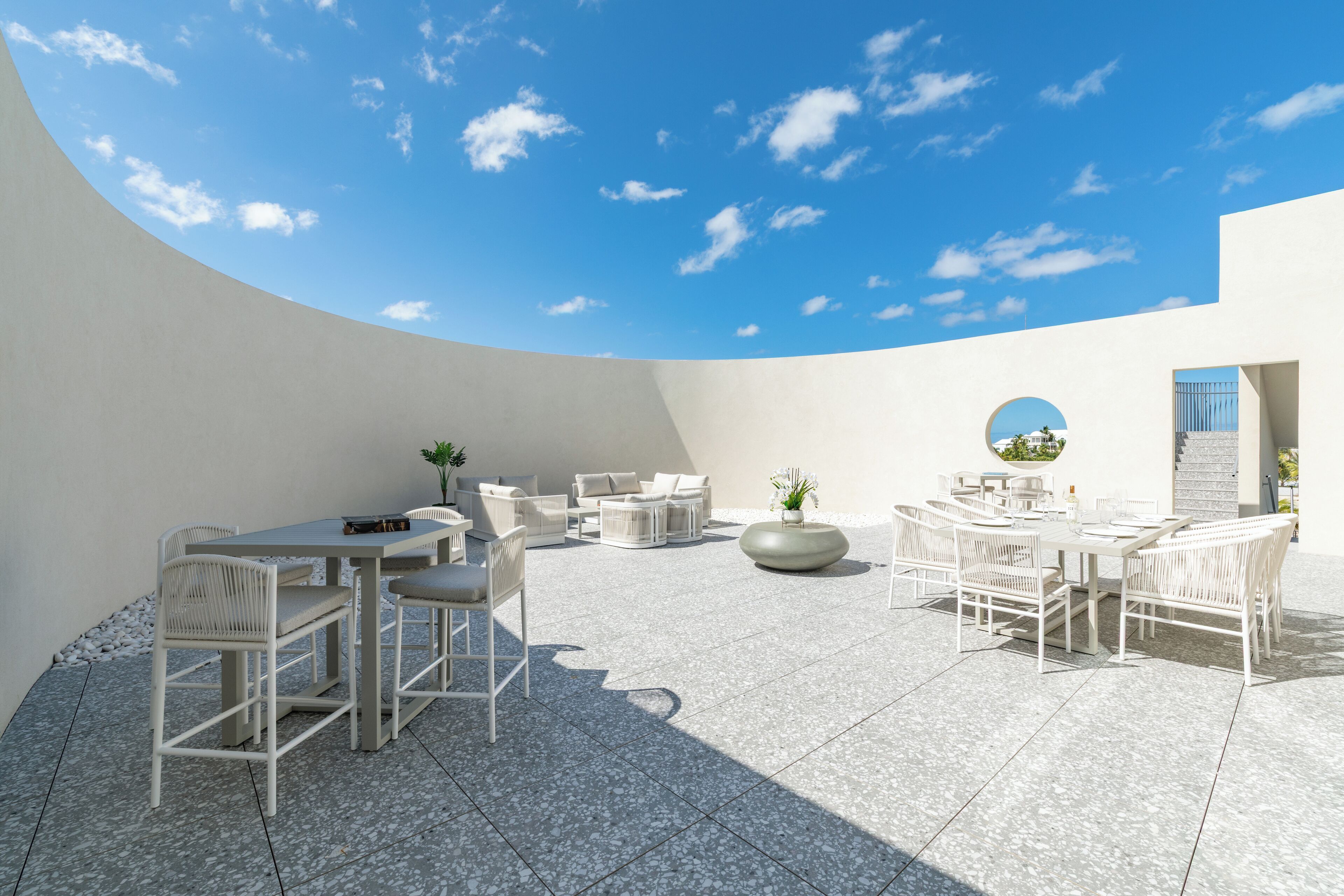 Um pátio externo com um muro branco curvo, uma mesa redonda com cadeiras e um espaço de estar com almofadas.