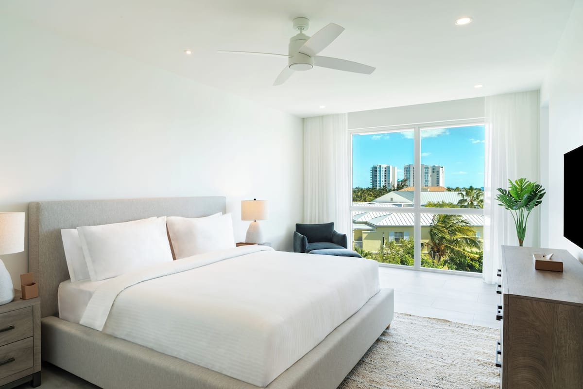 A modern hotel room with a large bed, a ceiling fan, a desk with a TV, and a view of buildings through large windows.