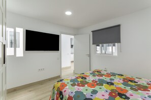 Ein Schlafzimmer mit einem Bett, einem Fernseher, einem Fenster mit Jalousie und einer Tür zu einem anderen Raum.