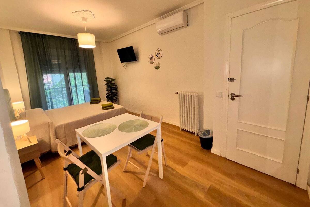 Een kleine, goed verlichte kamer met een bed, een tafel met stoelen, een radiator en een deur.