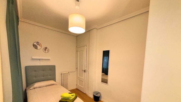 A bedroom with a bed, a radiator, a door, a window, and a trash bin. - Rooms Carlos, next to Plaza Mayor (Madrid)