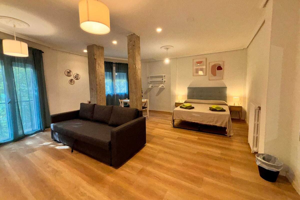 Een moderne woonkamer met een donkere bank, een bed met groene kussens, houten vloeren en hanglampen.