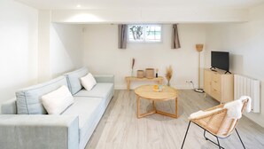 Una sala moderna con un sofá azul claro, una mesa de centro de madera, un sillón de mimbre, un televisor sobre un mueble bajo y una ventana con cortinas.
