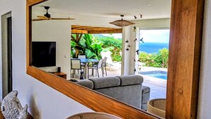 Un moderno salón con un espejo grande que refleja una piscina y vista al océano, un sofá gris y techo de madera.