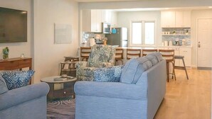 Un salon comprenant un canapé, des fauteuils, une table basse et un téléviseur. Au fond, on aperçoit une cuisine avec des placards et une fenêtre.