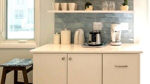 Une cuisine avec des meubles blancs, un plan de travail sur lequel se trouvent une machine à café, une bouilloire et un grille-pain. Il y a une fenêtre avec des stores et un petit tabouret.