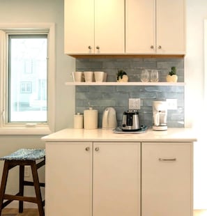 A kitchen with white cabinets, a countertop with a coffee maker, a kettle, and a toaster. There is a window with blinds and a small stool. - Charming Bethany Beach Getaway Pickle Ball (Bethany Beach)