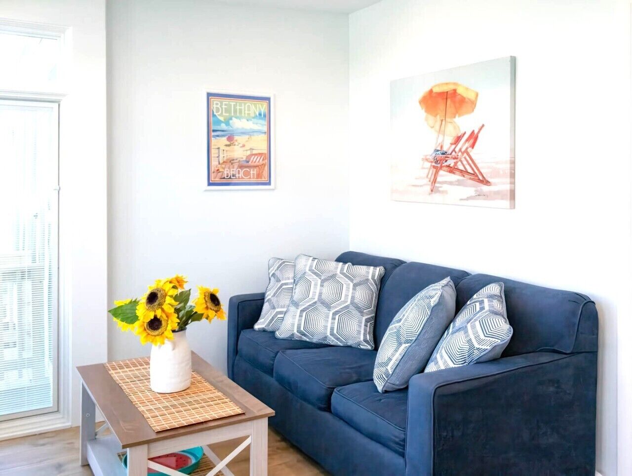 A living room with a blue sofa, patterned pillows, a vase with sunflowers, and two wall art pieces.