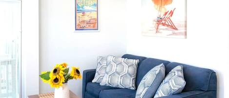 A living room with a blue sofa, patterned pillows, a vase with sunflowers, and two wall art pieces.