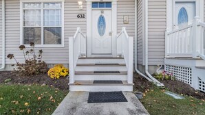 Une maison à deux étages avec une porte d’entrée blanche, le numéro « 632 » au-dessus de la porte, et un petit jardin devant la maison, agrémenté de fleurs jaunes et d’un abreuvoir pour oiseaux.