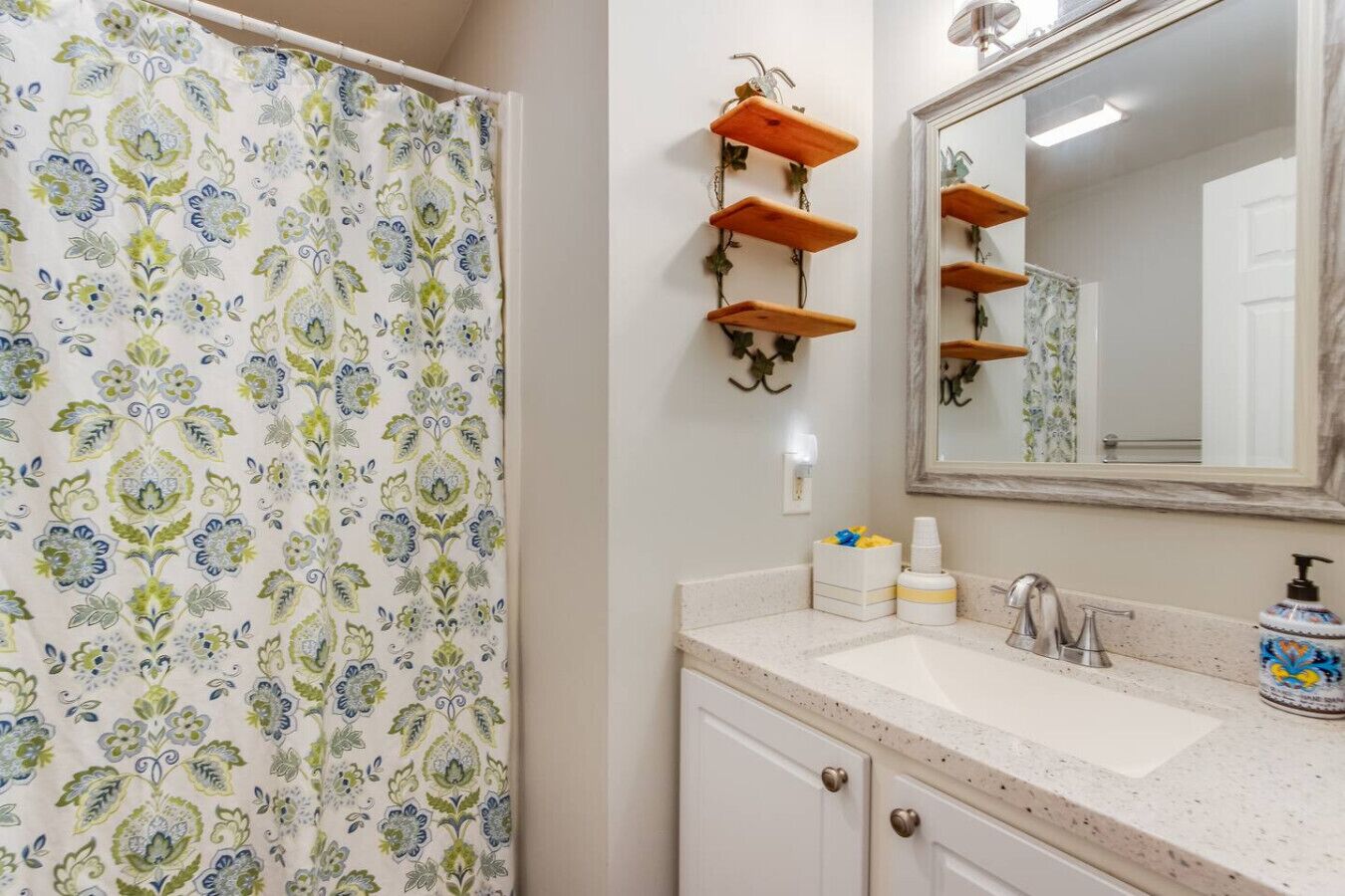 Une salle de bain avec un rideau de douche à motifs, un miroir surmonté d’une étagère en bois et un lavabo avec des articles de toilette.
