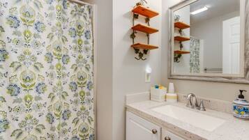 Une salle de bain avec un rideau de douche à motifs, un miroir surmonté d’une étagère en bois et un lavabo avec des articles de toilette.