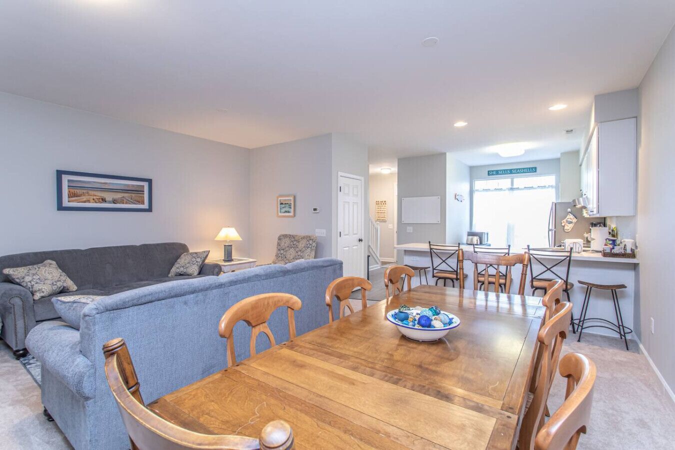 A modern living room with a grey sofa, wooden dining table, and chairs. A kitchen area is visible in the background with a sign reading "SHE SELLS SEASHELLS".