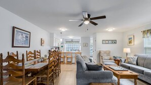 Un vaste salon comprenant un coin repas, un canapé gris, des chaises en bois, un ventilateur de plafond et, au fond, une cuisine.