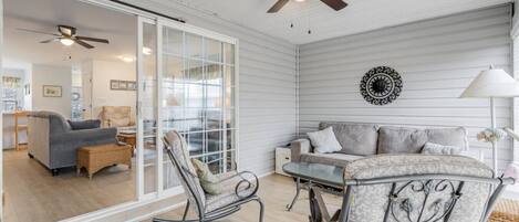 A spacious room with a sliding glass door leading to a balcony, a ceiling fan, a patterned armchair, a sofa, and a decorative wall piece.