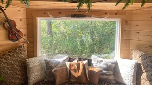 Interior acogedor de una cabaña con techo de madera, un violín colgado en la pared, una ventana con vista al bosque y una cama con almohadones estampados.