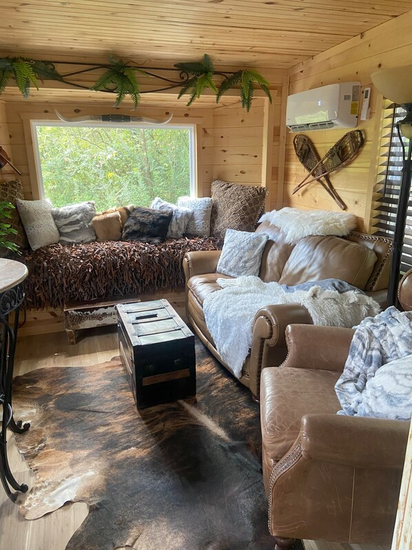 A cozy cabin interior with a wooden ceiling, a large window with a view of greenery, a leather sofa, a small table, and a rustic bench. -  Tiny Mountain Lodge with Smokey Mountain views. Newport TN Lodge decor. (Newport)