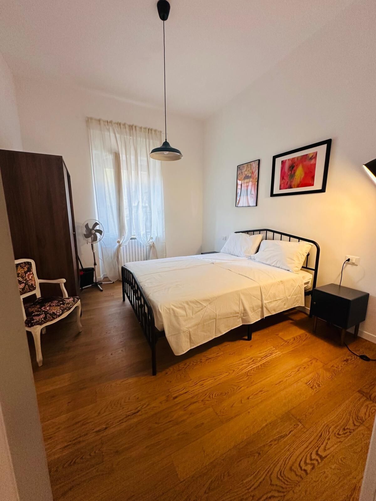 Un dormitorio con una cama, una silla, una mesita de noche, un ventilador y una ventana con cortinas.