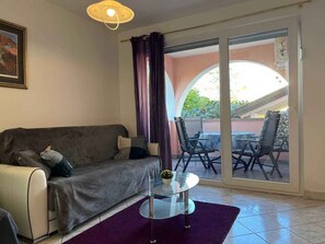A living room with a sofa, a glass coffee table, a purple rug, and a view of an outdoor patio with chairs and a table.
