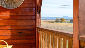 A wooden cabin with a woven basket hanging on the wall, a wooden railing, and a view of a rural landscape with fields and distant mountains.