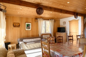 Interior de una cabaña de madera con sofá, mesa de comedor, sillas, televisor y ventana con cortinas.