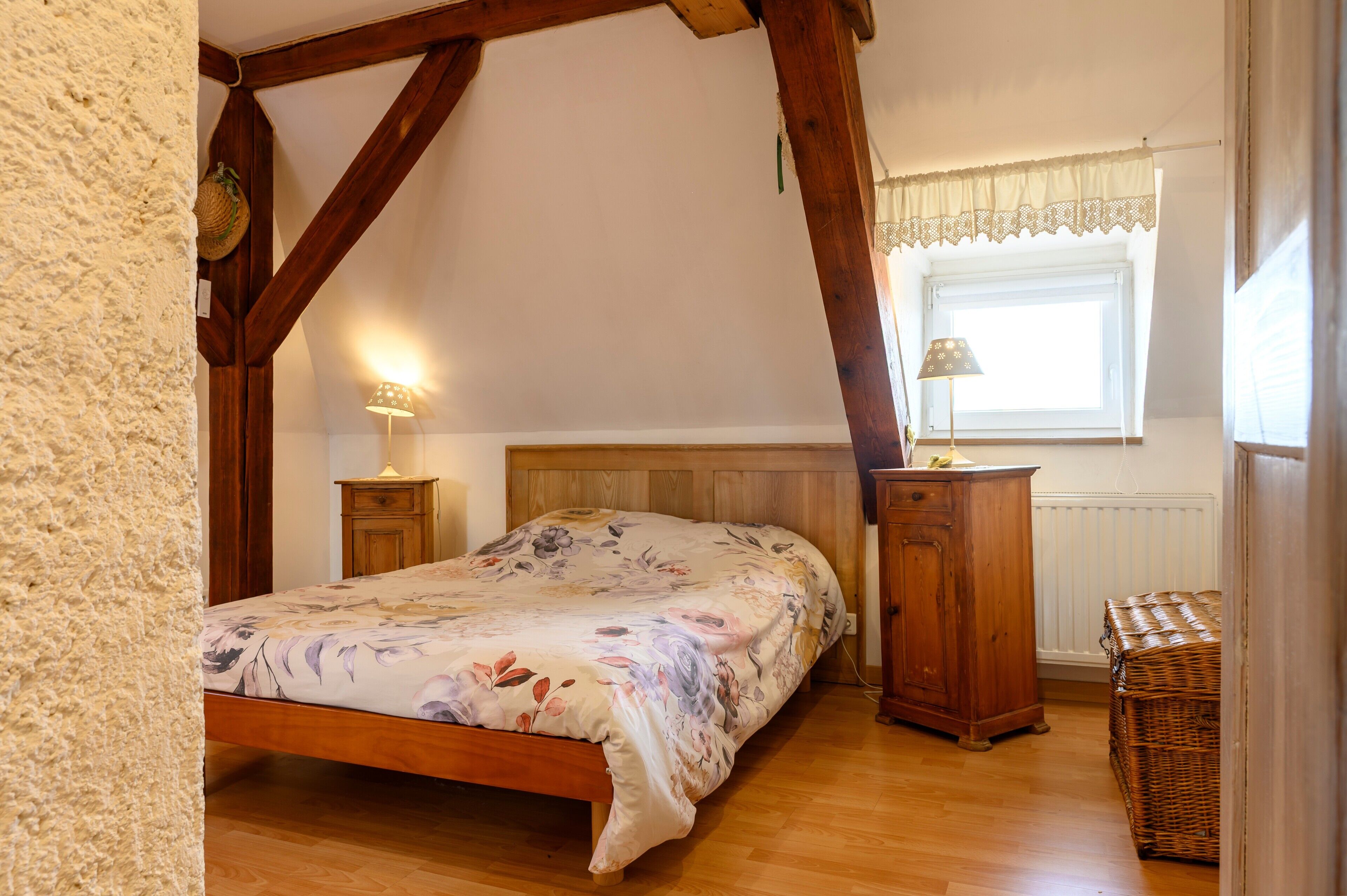 Un dormitorio con una cama de madera, una mesita de noche, un aparador, una cesta de mimbre y una ventana con un encaje blanco.