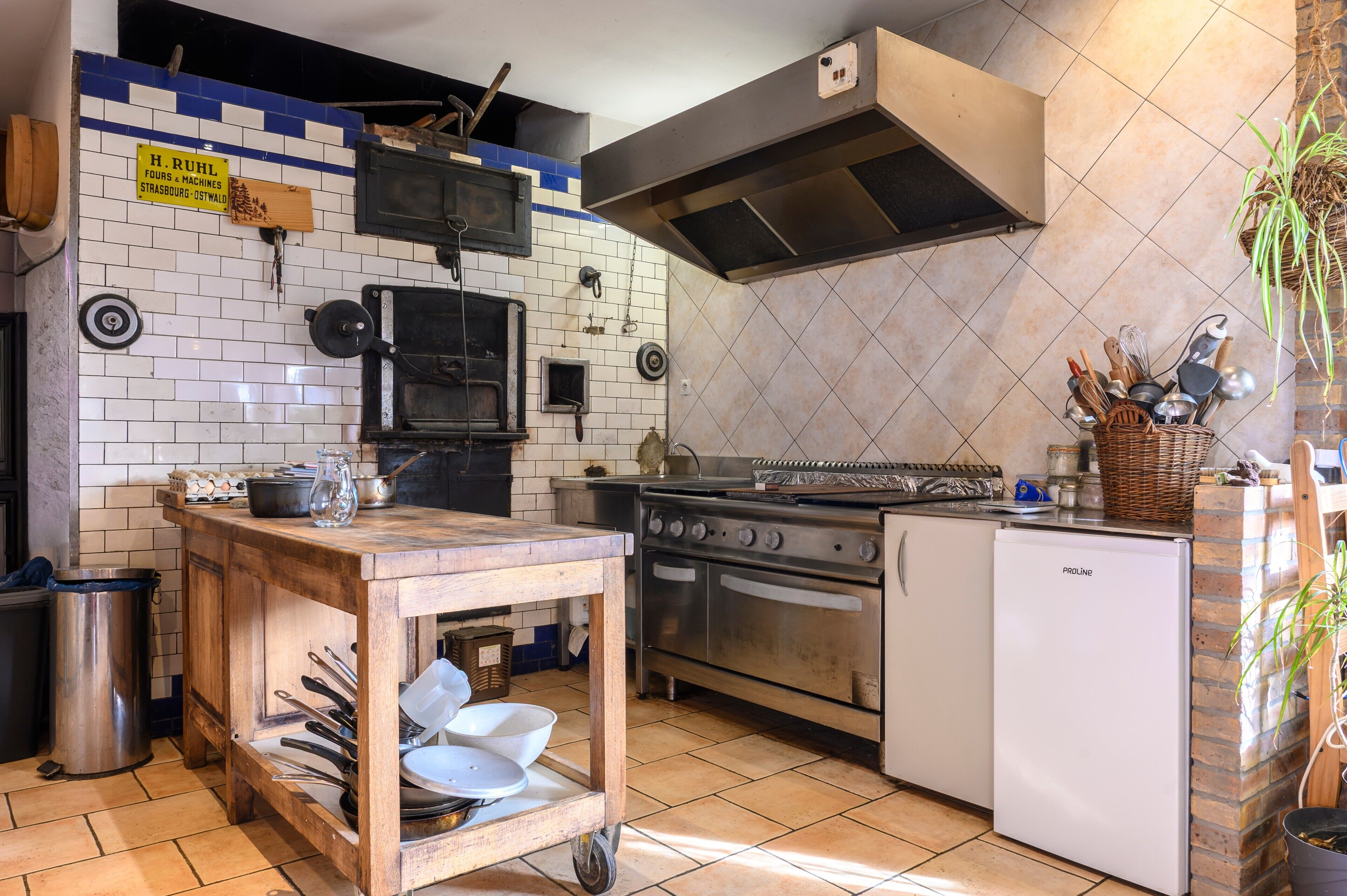 A kitchen with a stove, oven, and refrigerator, a wooden table with dishes, and a trash can.