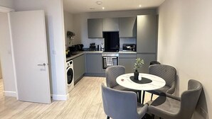 A modern kitchen with grey cabinets, a built-in oven, and a dining area with a round table and grey chairs. - Salford City Retreat - 2 Bedroom Apartment (Salford)