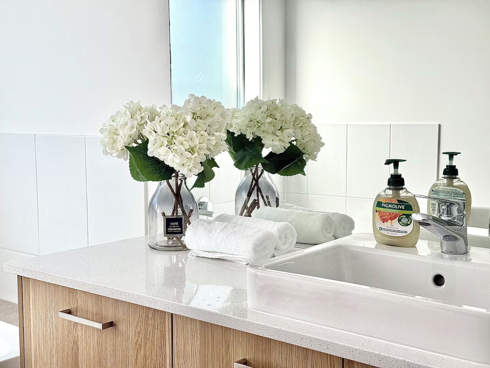A bathroom with a white sink, two bottles of Palmolive soap, a vase with white flowers, and rolled towels.
