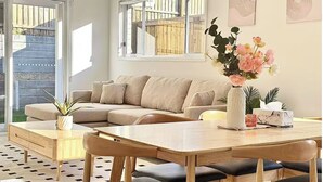 Ein modernes Wohnzimmer mit einem beigen Sofa, einem Holzesstisch mit schwarzen Stühlen, einer Blumenvase und Blick auf eine Holzterrasse durch große Fenster.