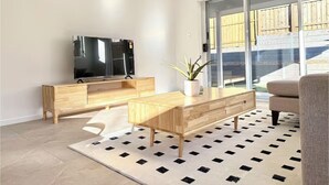 Ein modernes Wohnzimmer mit Flachbildfernseher, Holzmöbeln, einer Topfpflanze und Blick auf eine Holzdiele durch Glastüren.
