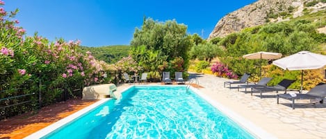 A swimming pool with lounge chairs and umbrellas, surrounded by greenery and a mountainous backdrop.