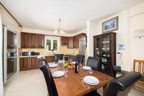A dining area with a wooden table set for four, surrounded by dark chairs. A kitchen with wooden cabinets and a refrigerator is visible in the background.