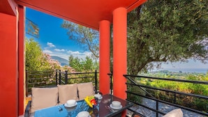 Ein Balkon mit Tisch für zwei Personen, umgeben von roten Wänden und Blick auf Grünflächen und Hügel.
