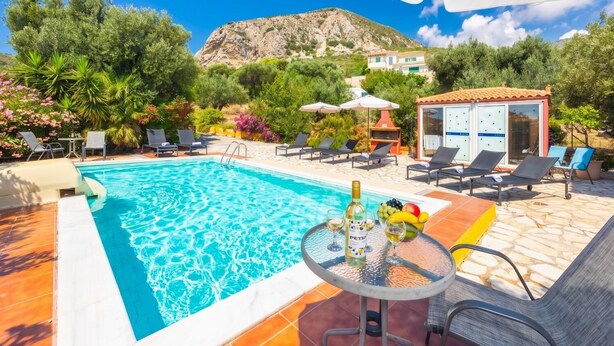 Ein Swimmingpool mit Liegestühlen und einem Tisch mit Getränken, umgeben von viel Grün und einem Berg im Hintergrund.