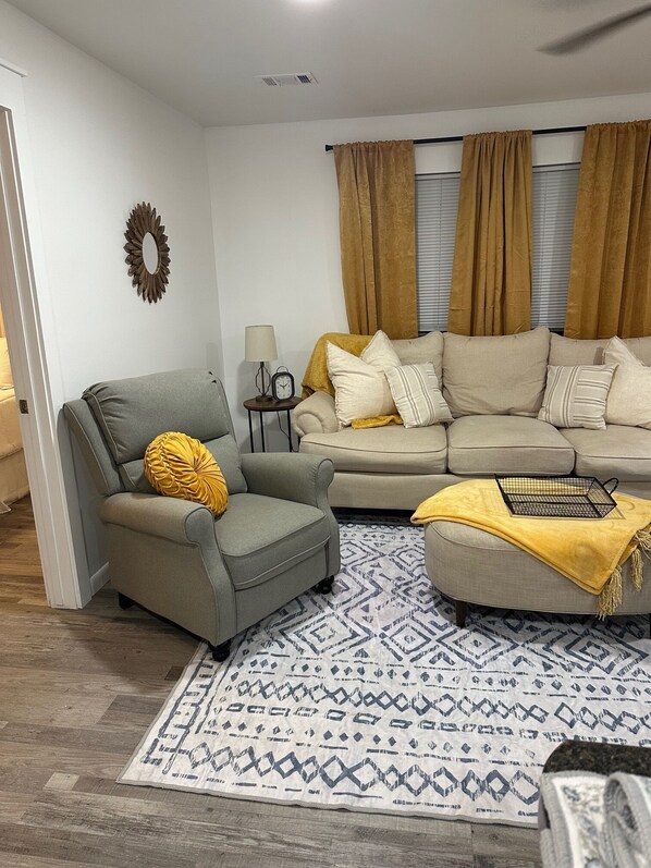 A living room with a grey armchair, a beige sofa, a patterned rug, and a wall-mounted mirror.