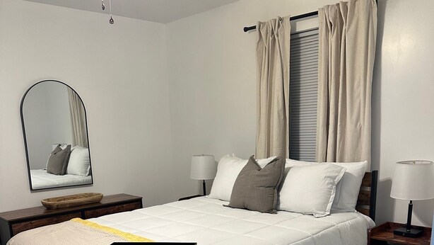 A bedroom with a bed, a nightstand, a mirror, a ceiling fan, and a window with curtains. - Canary Cottage in Beautiful Ailey, GA (Ailey)