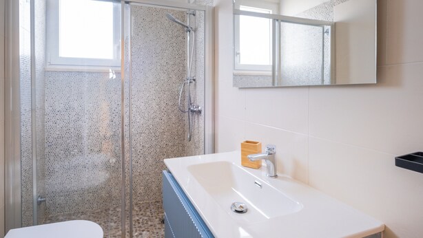 Ein modernes Badezimmer mit einer Duschkabine aus Glas, einem weißen Waschbecken mit einem hölzernen Seifenspender und einem großen Spiegel darüber.