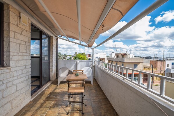 Un balcone con tavolo e sedie, vista sui palazzi e cielo sereno.
