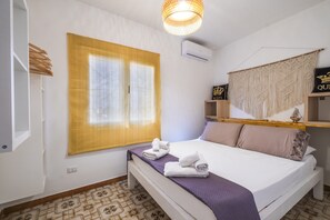 Una camera d'albergo con un letto, una finestra con tende gialle, un arazzo macramè e un piccolo ripiano con un cartello.