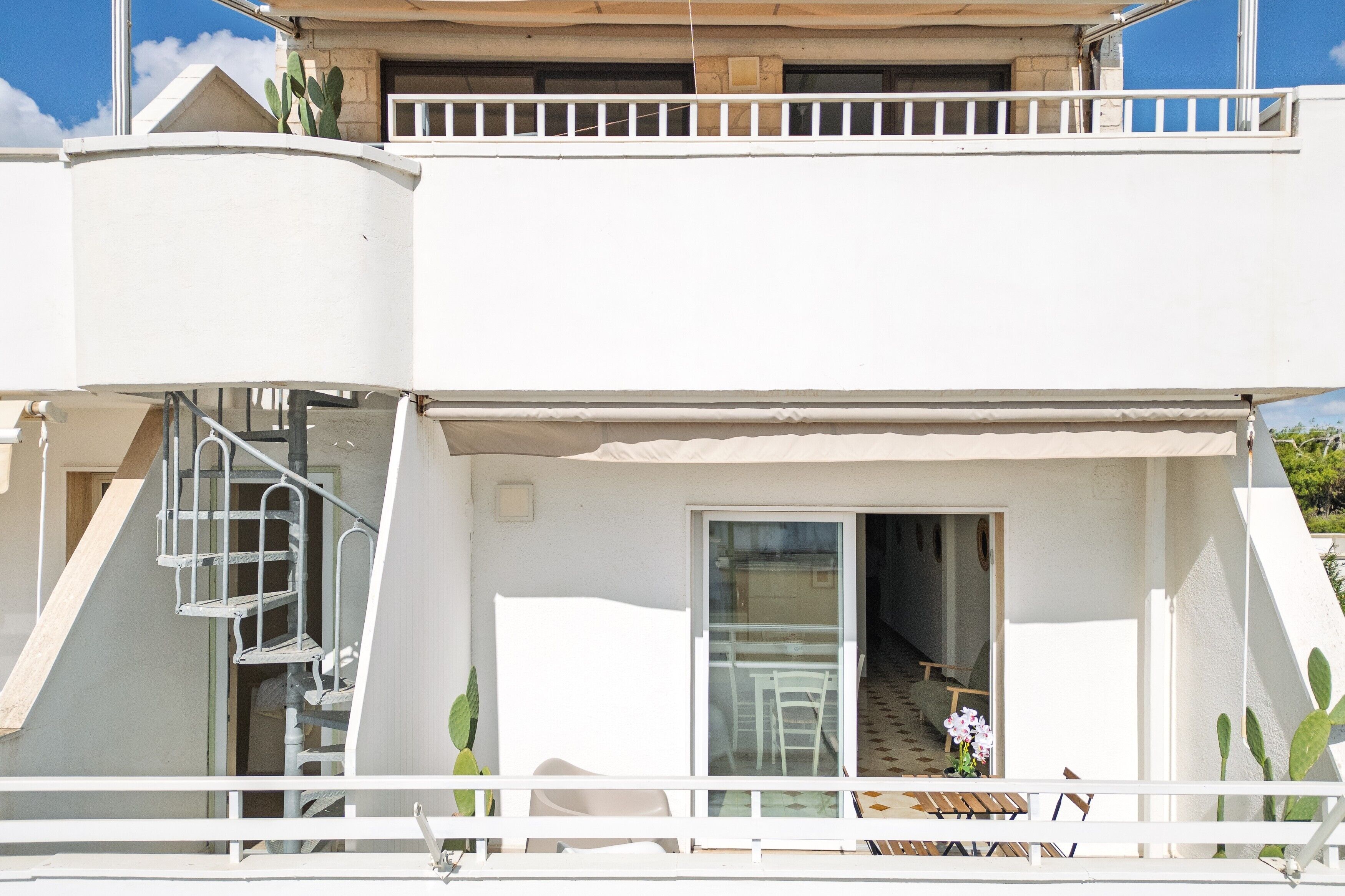 Un edificio a due piani con un balcone, una scala e un'ampia porta scorrevole che dà su un'area salotto all'aperto.