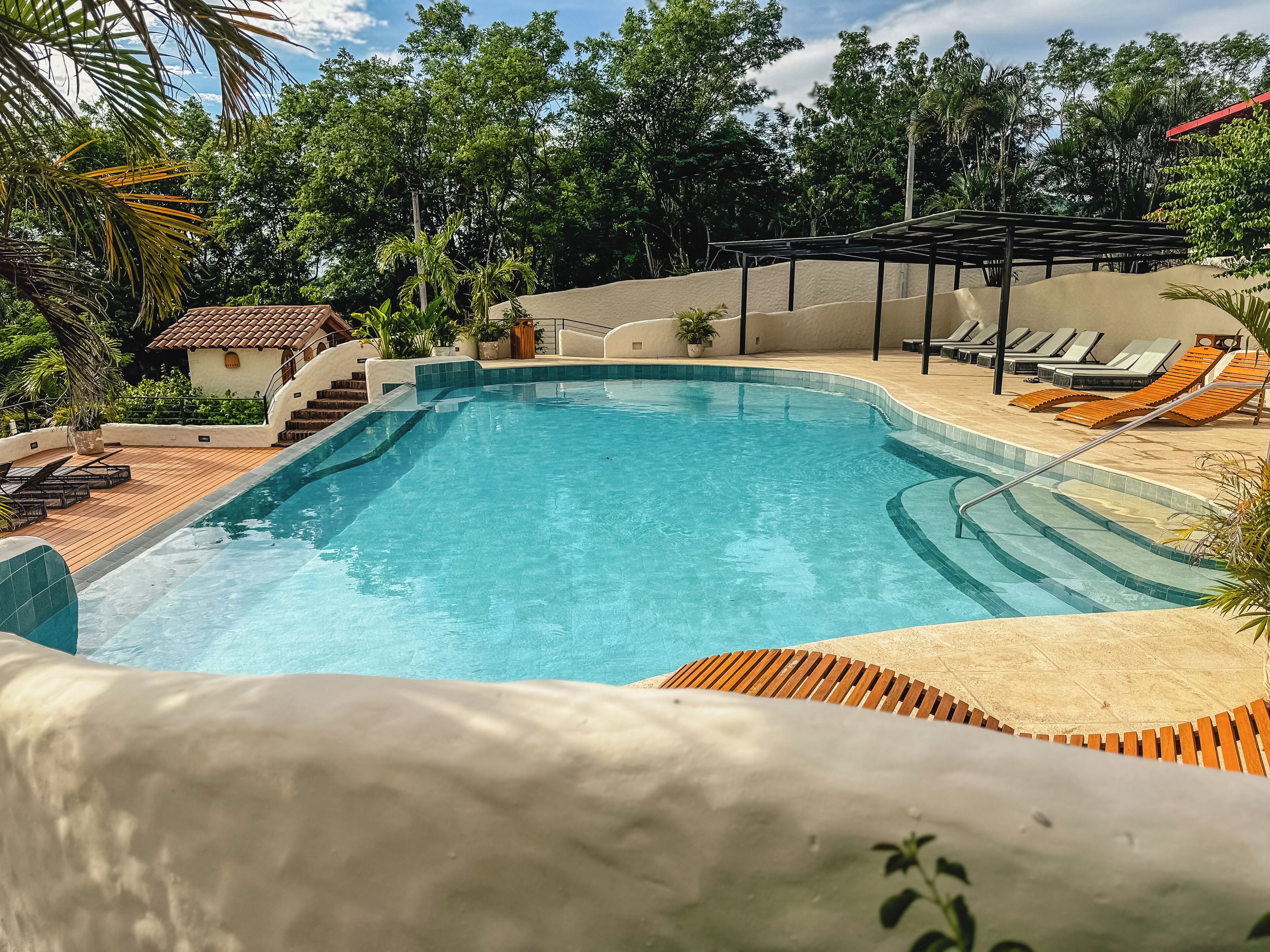 A swimming pool with a wooden deck, lounge chairs, and a covered area.