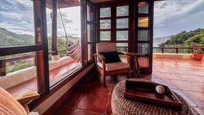 Un balcon spacieux avec une chaise en bois et un hamac, de grandes fenêtres offrant une vue sur les collines.