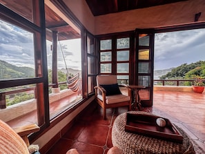 Un balcon spacieux avec une chaise en bois et un hamac, de grandes fenêtres offrant une vue sur les collines.